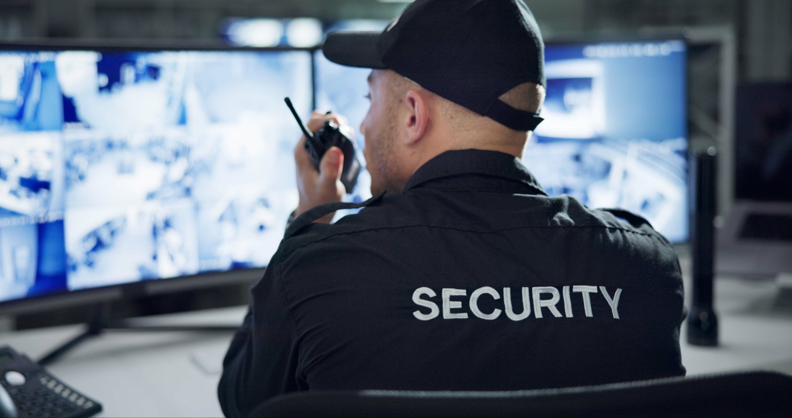 Male security guard, radio and speaking by cctv monitor for protection, data center or building safety. Inspection, video surveillance agency and control room with footage, back view and computer Security personnel monitoring surveillance cameras at a control center, using a walkie-talkie to communicate