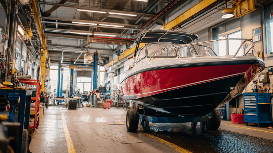 A red motorboat inside a spacious repair shop, surrounded by tools, workbenches, and industrial equipment.