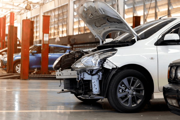 White car with front-end damage and hood open inside an auto repair shop, with other vehicles and service bays in the background.