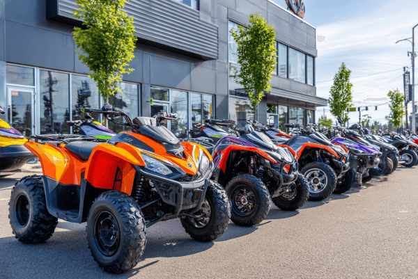 A row of brightly colored ATVs and motorcycles lined up outside a modern powersports dealership, showcasing inventory on the lot under clear skies.
