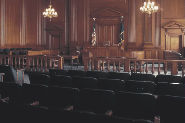 Empty courtroom with wooden walls, judge’s bench, witness stand, and rows of dark seats for the jury and audience. American and state flags stand behind the judge’s bench under a sign reading ‘In God We Trust.